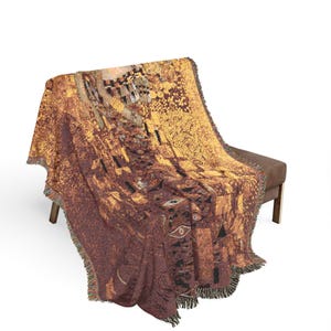 Puede incluir: Una manta tejida con un diseño abstracto detallado en tonos marrón, dorado y negro. La manta está sobre un banco marrón, mostrando sus bordes con flecos y su intrincado patrón. El diseño presenta formas geométricas y motivos de ojos.