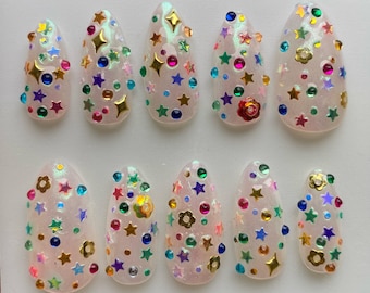 Puede incluir: Un conjunto de diez uñas artificiales, cada una con una base nacarada y adornada con adornos coloridos en forma de estrella, flor y gema. Las decoraciones incluyen acentos dorados, azules, verdes, rosas y morados.