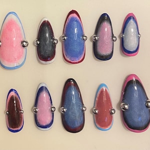 Pode incluir: Um conjunto de dez unhas artificiais com vários designs. Cada unha em forma de amêndoa apresenta um gradiente de cores, com tons de rosa, azul, castanho e preto. Tachas metálicas prateadas são fixadas em cada unha.