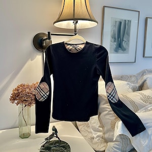 Peut inclure: Haut noir à manches longues avec des coudières et un motif à carreaux. Le chemisier est suspendu à un cintre. Le haut a un col rond et le motif à carreaux est également visible à l'intérieur du col.