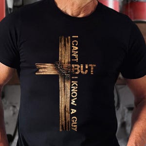 May include: Black t-shirt featuring a wooden cross design with the text "I CAN'T BUT I KNOW A GUY" in a rustic font. The cross is made of wooden planks and has a metal crown of thorns.