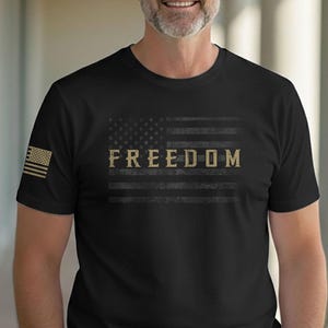 May include: Black t-shirt featuring a distressed American flag design with the word "FREEDOM" in a gold color. The shirt has a crew neck and short sleeves. A small American flag is on the left sleeve.