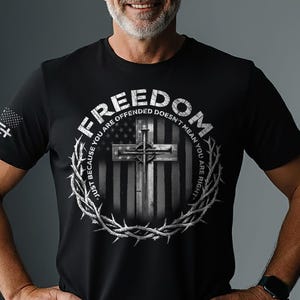 May include: Black t-shirt featuring a graphic design with a cross, the American flag, and the word "FREEDOM". The design is encircled by a crown of thorns. The text "JUST BECAUSE YOU ARE OFFENDED DOESN'T MEAN YOU ARE RIGHT" is also included.