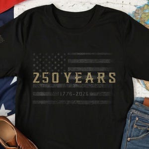 K&ouml;nnte beinhalten: Schwarzes T-Shirt mit einem verblassten amerikanischen Flaggen-Design und dem Text "250 YEARS 1776-2026". Das T-Shirt hat einen Rundhalsausschnitt und kurze &Auml;rmel. Das Shirt wird mit braunen Lederschuhen, Jeans und einer amerikanischen Flagge pr&auml;sentiert.
