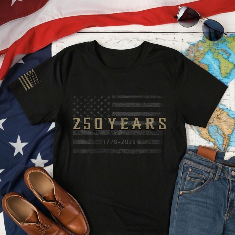 K&ouml;nnte beinhalten: Schwarzes T-Shirt mit einem verblassten amerikanischen Flaggen-Design und dem Text "250 YEARS 1776-2026". Das T-Shirt hat einen Rundhalsausschnitt und kurze &Auml;rmel. Das Shirt wird mit braunen Lederschuhen, Jeans und einer amerikanischen Flagge pr&auml;sentiert.