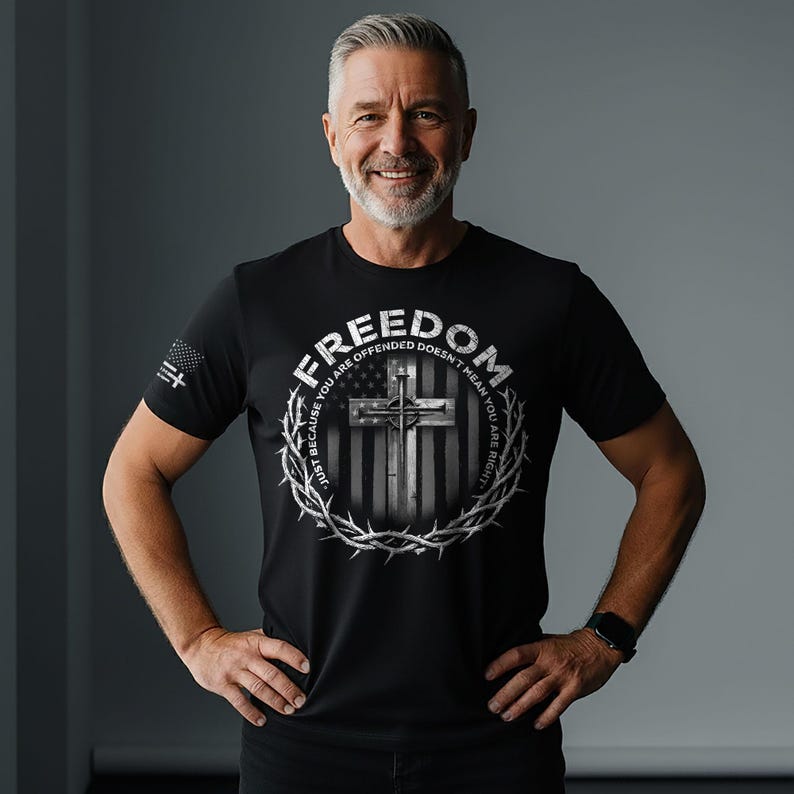 May include: Black t-shirt featuring a graphic design with a cross, the American flag, and the word "FREEDOM". The design is encircled by a crown of thorns. The text "JUST BECAUSE YOU ARE OFFENDED DOESN'T MEAN YOU ARE RIGHT" is also included.