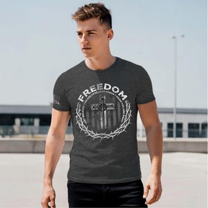 May include: Dark gray t-shirt with the word "FREEDOM" in a circular design. The design includes a cross, barbed wire, and the text "JUST BECAUSE YOU ARE OFFENDED DOESN'T MEAN YOU ARE RIGHT." The shirt has a small American flag on the sleeve.