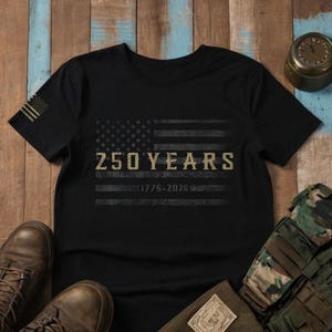 K&ouml;nnte beinhalten: Schwarzes T-Shirt mit einem verblassten Design der amerikanischen Flagge und dem Text "250 YEARS 1776-2026". Das T-Shirt hat eine kleine amerikanische Flagge auf dem linken &Auml;rmel. Das T-Shirt wird mit Stiefeln und Milit&auml;rausr&uuml;stung pr&auml;sentiert.