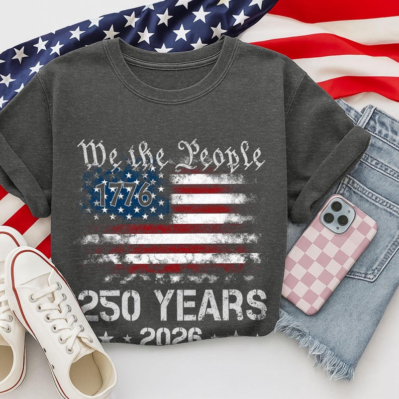May include: Dark gray t-shirt featuring an American flag design with the text "We the People 1776" and "250 YEARS 2026". The shirt is paired with blue denim shorts, white sneakers, and a pink checkered phone case.