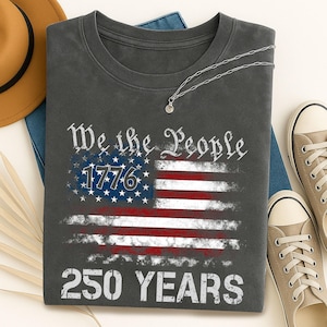 May include: Dark gray t-shirt featuring a distressed American flag design with the text "We the People 1776 250 YEARS." The shirt is paired with a silver necklace, tan sneakers, and a brown hat.