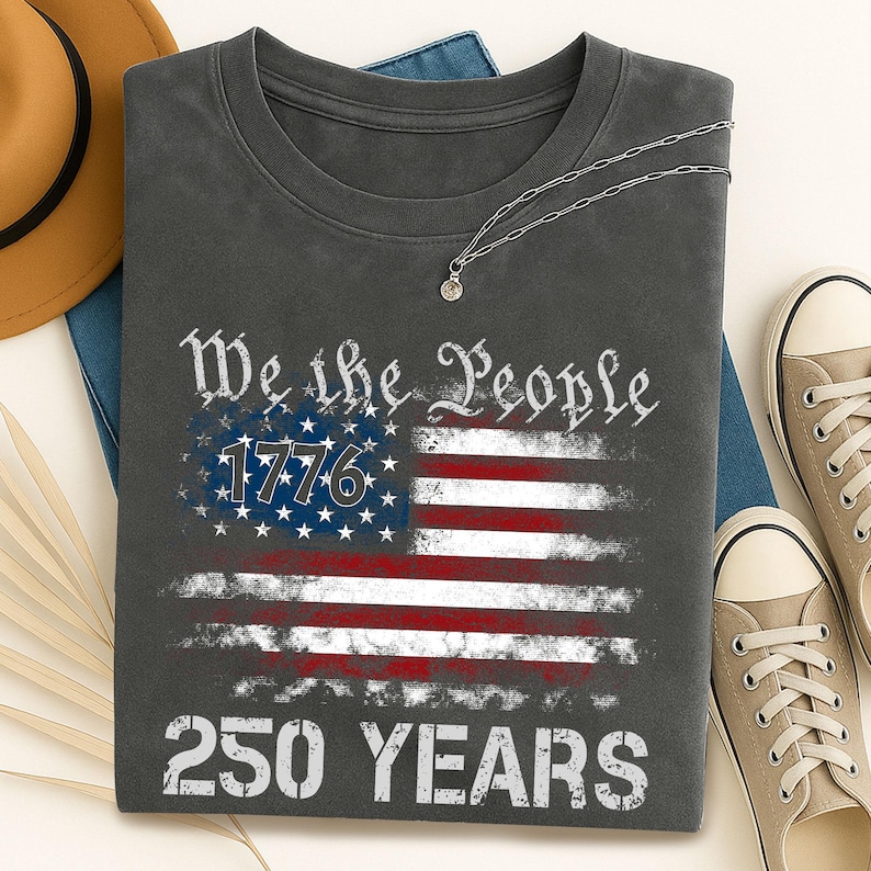 May include: Dark gray t-shirt featuring a distressed American flag design with the text "We the People 1776 250 YEARS." The shirt is paired with a silver necklace, tan sneakers, and a brown hat.