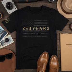 K&ouml;nnte beinhalten: Schwarzes T-Shirt mit einem verblassten amerikanischen Flaggen-Design und dem goldenen Text "250 YEARS 1776-2026". Das T-Shirt liegt auf einer Holzoberfl&auml;che mit einer Kamera, Sonnenbrille und braunen Lederschuhen. Das Design feiert ein Jubil&auml;um.
