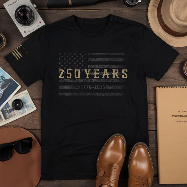 K&ouml;nnte beinhalten: Schwarzes T-Shirt mit einem verblassten amerikanischen Flaggen-Design und dem goldenen Text "250 YEARS 1776-2026". Das T-Shirt liegt auf einer Holzoberfl&auml;che mit einer Kamera, Sonnenbrille und braunen Lederschuhen. Das Design feiert ein Jubil&auml;um.