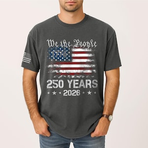 May include: Dark gray t-shirt featuring a distressed American flag design. The flag includes the text "We the People," "1776," "250 YEARS," and "2026." The shirt has a small American flag on the left sleeve.