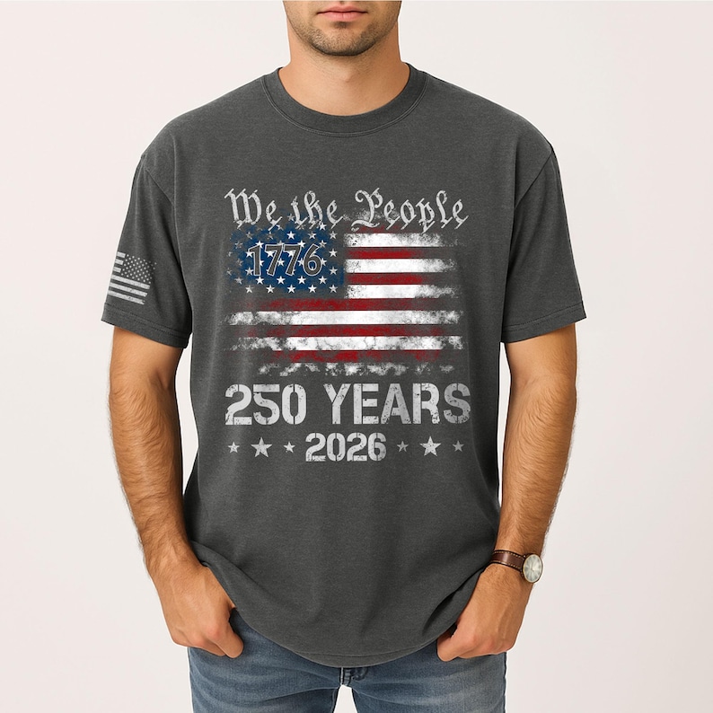 May include: Dark gray t-shirt featuring a distressed American flag design. The flag includes the text "We the People," "1776," "250 YEARS," and "2026." The shirt has a small American flag on the left sleeve.