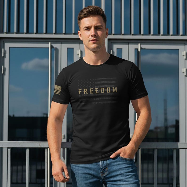 May include: Black crew neck t-shirt with a distressed American flag design and the word "FREEDOM" in gold lettering. The shirt has a small American flag on the left sleeve. The model is wearing blue jeans.