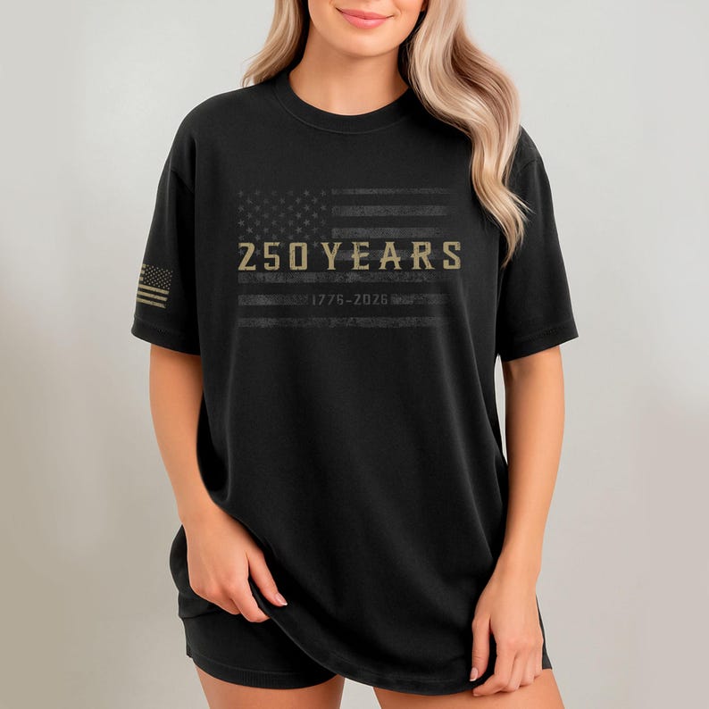 K&ouml;nnte beinhalten: Schwarzes T-Shirt mit einem verblassten Design der amerikanischen Flagge. Das Shirt zeigt den Text "250 YEARS" &uuml;ber "1776-2026" in Gold. Auf dem linken &Auml;rmel befindet sich eine kleine amerikanische Flagge.