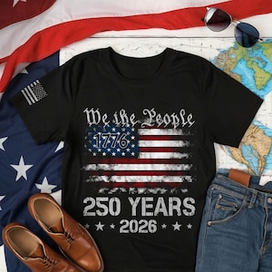 May include: Black t-shirt featuring an American flag design with the text "We the People 1776" and "250 YEARS 2026". The shirt also has a small American flag design on the left sleeve. The shirt is displayed with a pair of brown leather shoes and blue jeans.