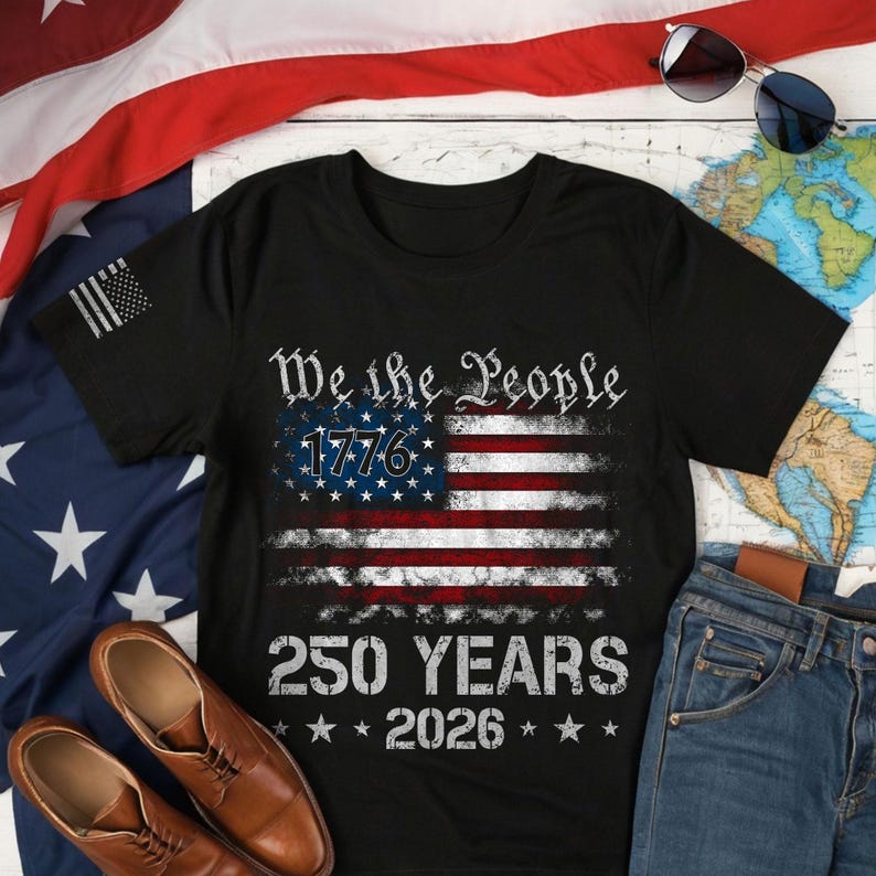 May include: Black t-shirt featuring an American flag design with the text "We the People 1776" and "250 YEARS 2026". The shirt also has a small American flag design on the left sleeve. The shirt is displayed with a pair of brown leather shoes and blue jeans.