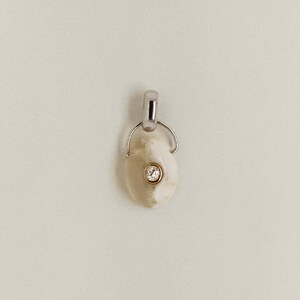 Puede incluir: Un colgante con una piedra ovalada de color crema y una peque&ntilde;a gema redonda y transparente engastada en un bisel dorado. El colgante tiene una fianza plateada para sujetarlo a una cadena. La joya est&aacute; sobre un fondo neutro.