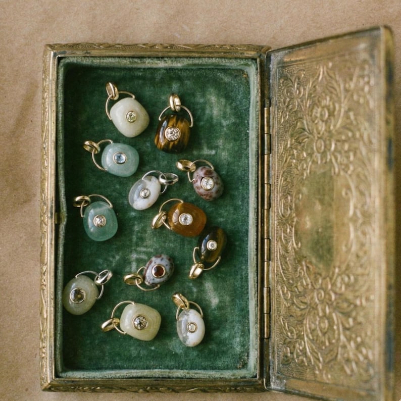 Puede incluir: Una colecci&oacute;n de colgantes de piedras preciosas exhibidos en una caja de joyer&iacute;a dorada antigua abierta, con un interior de terciopelo verde. Cada colgante presenta una piedra &uacute;nica con una fianza dorada y un peque&ntilde;o acento brillante.