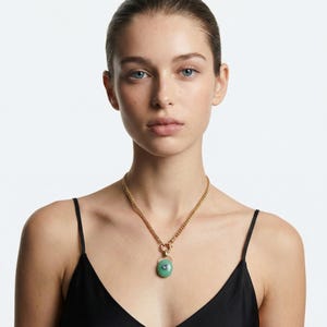 Puede incluir: Un collar de cadena dorada con un colgante redondo verde claro. El colgante tiene una peque&ntilde;a piedra azul claro en el centro. La mujer de la imagen lleva un top negro con tirantes finos.