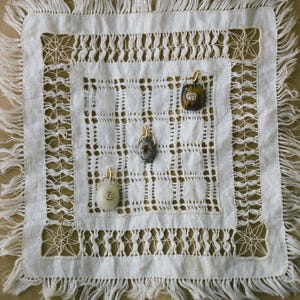 Puede incluir: Cuatro collares con colgantes con aros dorados se exhiben sobre un mantel individual cuadrado blanco de tejido abierto con flecos. Los colgantes tienen varios dise&ntilde;os de piedras y gemas.