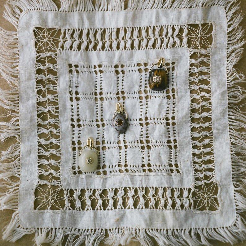 Puede incluir: Cuatro collares con colgantes con aros dorados se exhiben sobre un mantel individual cuadrado blanco de tejido abierto con flecos. Los colgantes tienen varios dise&ntilde;os de piedras y gemas.