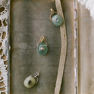 Puede incluir: Tres colgantes de piedras preciosas con detalles dorados y plateados. Los colgantes ovalados son de color verde claro y presentan peque&ntilde;as piedras transparentes. Los colgantes se exhiben sobre una superficie de tono neutro, con una cinta decorativa y un ribete de encaje.