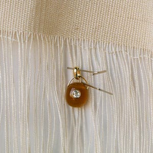 Puede incluir: Un peque&ntilde;o colgante dorado con una piedra marr&oacute;n de forma ovalada y una gema clara en el centro. El colgante est&aacute; sujeto a una tela blanca con flecos con un alfiler plateado. La piedra mide aproximadamente 1 cm de largo.