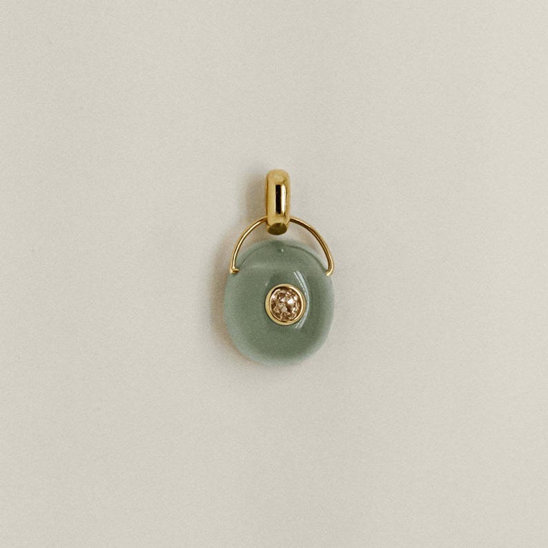 Puede incluir: Un colgante de oro con una piedra ovalada de color verde claro y una peque&ntilde;a piedra central redonda y brillante. El colgante tiene una fianza de oro y un lazo de oro que conecta con la piedra.