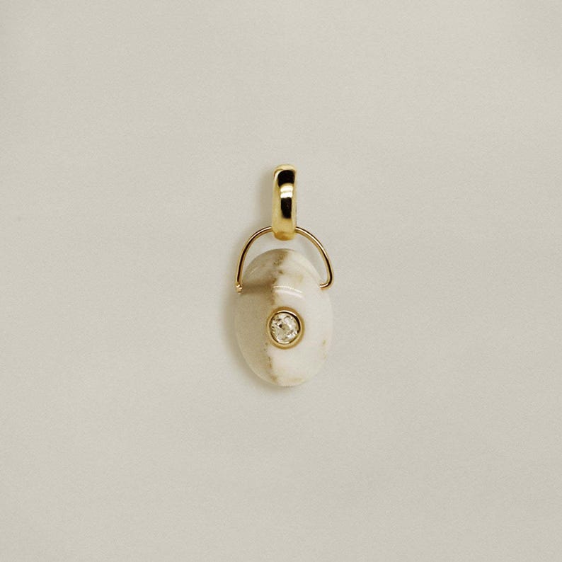 Puede incluir: Un colgante dorado con una piedra blanca y beige, con una peque&ntilde;a gema redonda y transparente en el centro. El colgante tiene un lazo en la parte superior para una cadena. El fondo es de color beige claro s&oacute;lido.