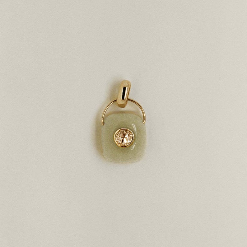 Puede incluir: Colgante de oro con una piedra cuadrada de color verde claro y una piedra redonda y transparente en el centro. El colgante tiene un lazo dorado en la parte superior para una cadena. El fondo es de color blanco roto.