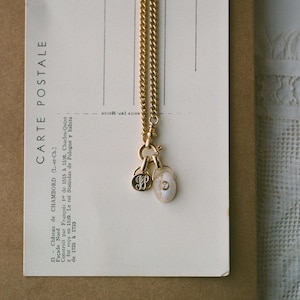 Puede incluir: Un collar dorado con una cadena fina y dos colgantes. Uno tiene las iniciales "JP", el otro es un colgante ovalado con un centro nacarado. El collar cuelga sobre una postal vintage con texto en franc&eacute;s.