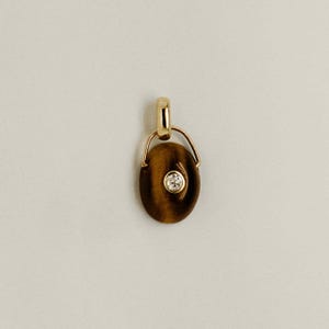 Puede incluir: Un colgante de oro con una piedra ojo de tigre ovalada y una peque&ntilde;a gema redonda y transparente en el centro. El colgante tiene un dise&ntilde;o sencillo y elegante con una fianza de oro para una cadena de collar. La piedra tiene tonos marrones y dorados.