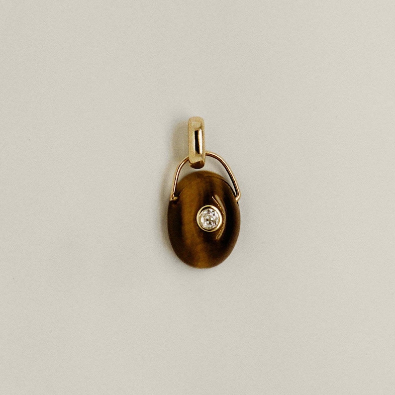Puede incluir: Un colgante de oro con una piedra ojo de tigre ovalada y una peque&ntilde;a gema redonda y transparente en el centro. El colgante tiene un dise&ntilde;o sencillo y elegante con una fianza de oro para una cadena de collar. La piedra tiene tonos marrones y dorados.