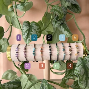 Può includere: Esposizione di nove braccialetti con pietre preziose, ognuno con una combinazione di colori e disposizione di pietre unica. I braccialetti sono infilati su cordini neri o marroni e sono esposti su un supporto cilindrico beige, circondati da piante verdi.