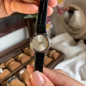 May include: A silver-toned Credor Seiko watch with a cream-colored face and black leather band. The watch has gold-colored hands and hour markers. The watch is held up in front of a jewelry box.