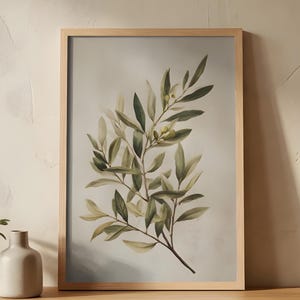 May include: A framed botanical print featuring an olive branch with green and yellow leaves. The artwork is displayed on a wooden table, alongside a small potted plant and a ceramic vase. The frame is made of light-colored wood.