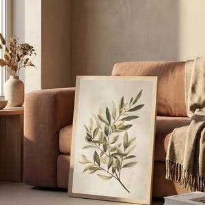 May include: A framed botanical print featuring a detailed illustration of an olive branch with green leaves and small yellow olives. The artwork is set against a soft, off-white background and framed in light wood.