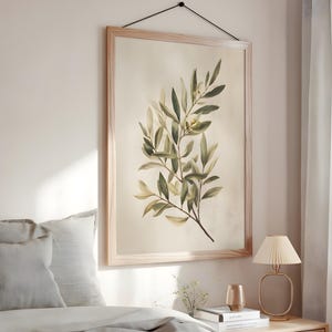 May include: A framed botanical print featuring a watercolor illustration of olive branches with green and yellow leaves. The artwork is displayed in a light wood frame and hangs on a beige wall. The print is suspended by a black cord.