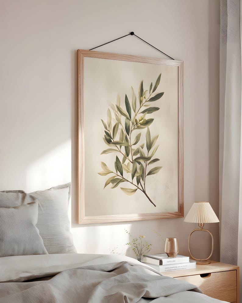 May include: A framed botanical print featuring a watercolor illustration of olive branches with green and yellow leaves. The artwork is displayed in a light wood frame and hangs on a beige wall. The print is suspended by a black cord.