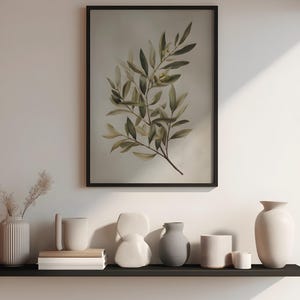 May include: A framed botanical print of an olive branch with green leaves and small olives. Below the print, a black shelf displays a collection of ceramic vases and decorative objects in neutral tones, including white, gray, and beige.