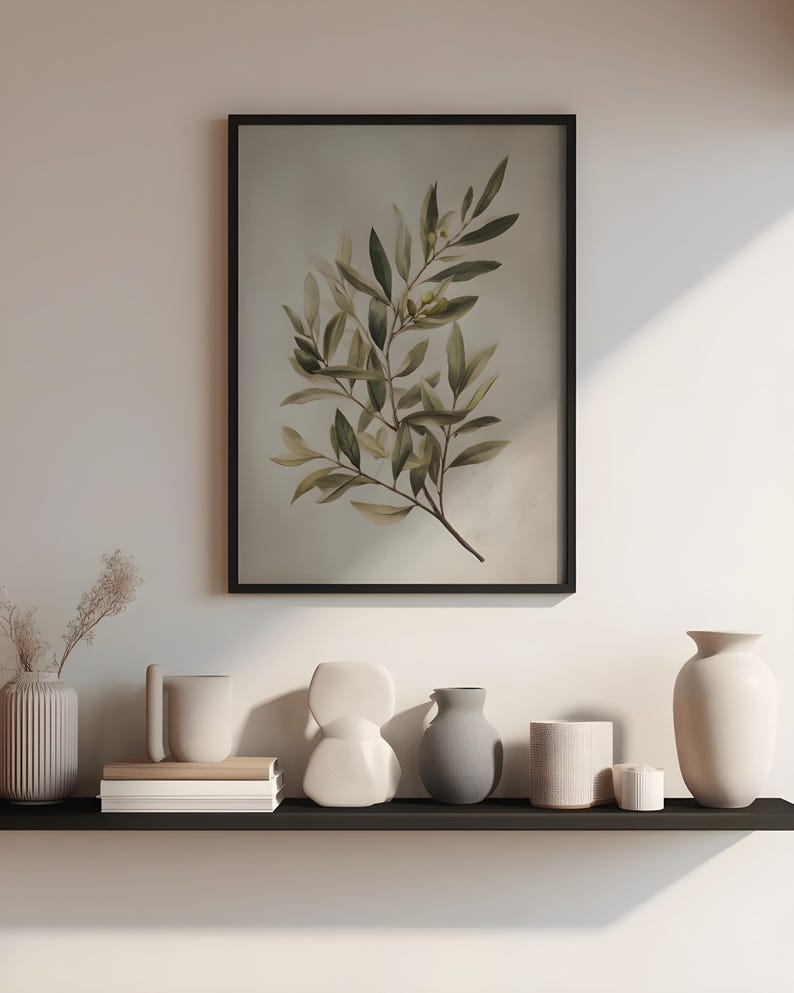 May include: A framed botanical print of an olive branch with green leaves and small olives. Below the print, a black shelf displays a collection of ceramic vases and decorative objects in neutral tones, including white, gray, and beige.
