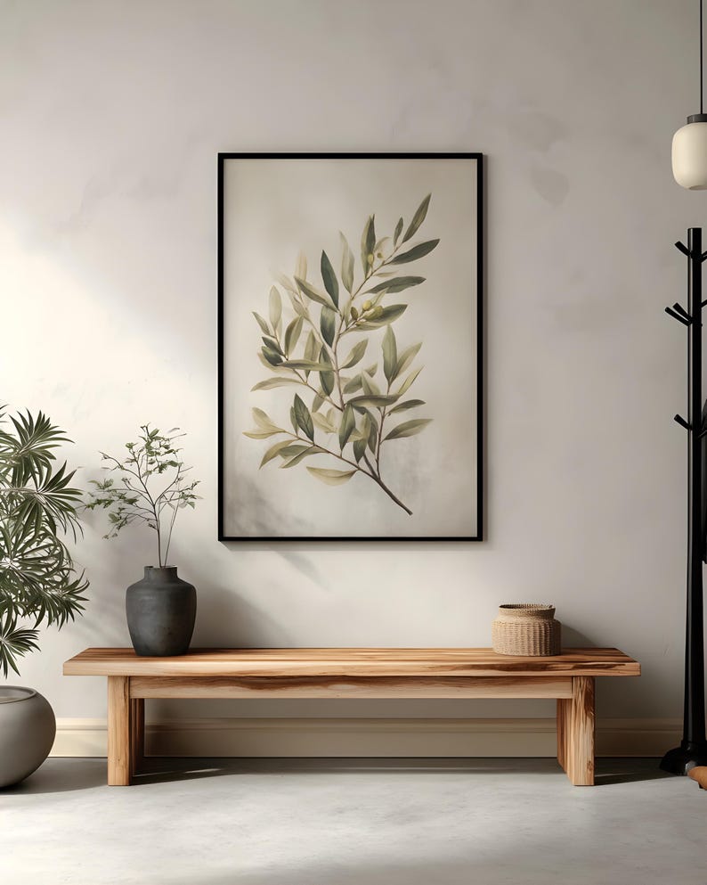 May include: A framed botanical print featuring a watercolor illustration of olive branches with green and yellow leaves. The artwork hangs above a wooden bench, with a dark vase and a woven basket adding to the decor. The room has a neutral color palette.