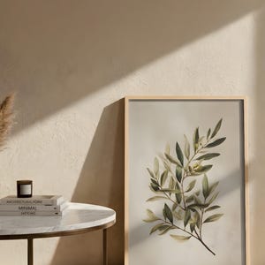 May include: A framed botanical print featuring a detailed illustration of an olive branch with green and yellow leaves. The artwork is set against a neutral background and framed in light wood. The print is displayed next to a small side table with books and a candle.
