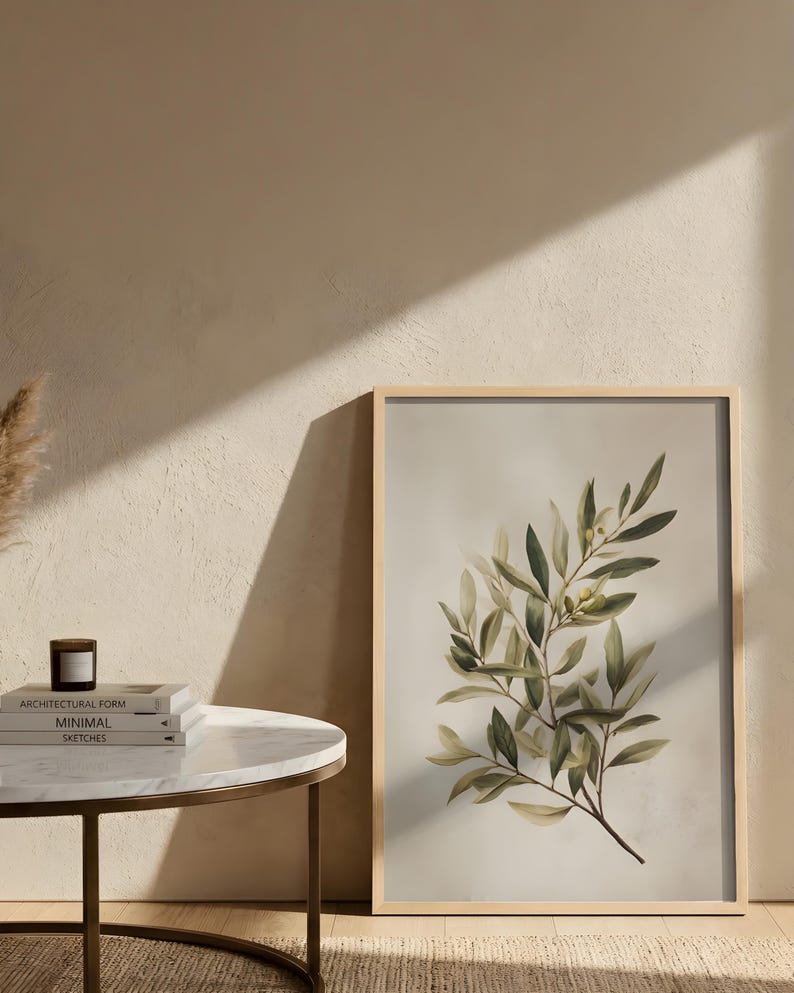 May include: A framed botanical print featuring a detailed illustration of an olive branch with green and yellow leaves. The artwork is set against a neutral background and framed in light wood. The print is displayed next to a small side table with books and a candle.