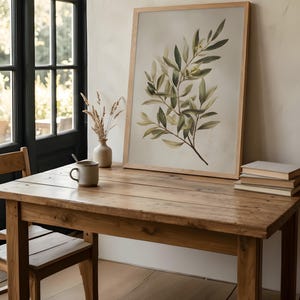 May include: A framed botanical print featuring a watercolor illustration of olive branches with green leaves and small olives. The artwork is displayed on a rustic wooden table, alongside a ceramic mug, vase, and a stack of books. The room has a neutral color palette.