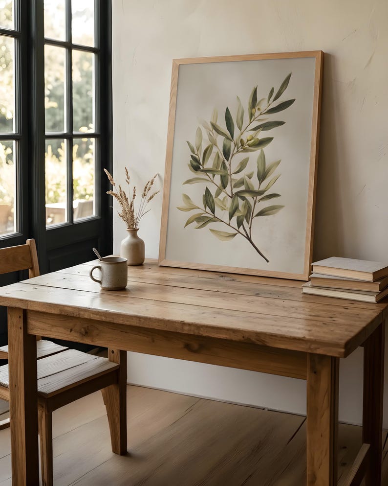 May include: A framed botanical print featuring a watercolor illustration of olive branches with green leaves and small olives. The artwork is displayed on a rustic wooden table, alongside a ceramic mug, vase, and a stack of books. The room has a neutral color palette.