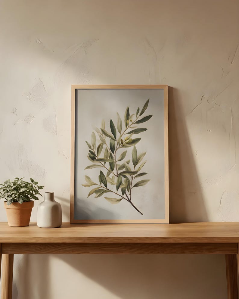May include: A framed botanical print featuring an olive branch with green and yellow leaves. The artwork is displayed on a wooden table, alongside a small potted plant and a ceramic vase. The frame is made of light-colored wood.